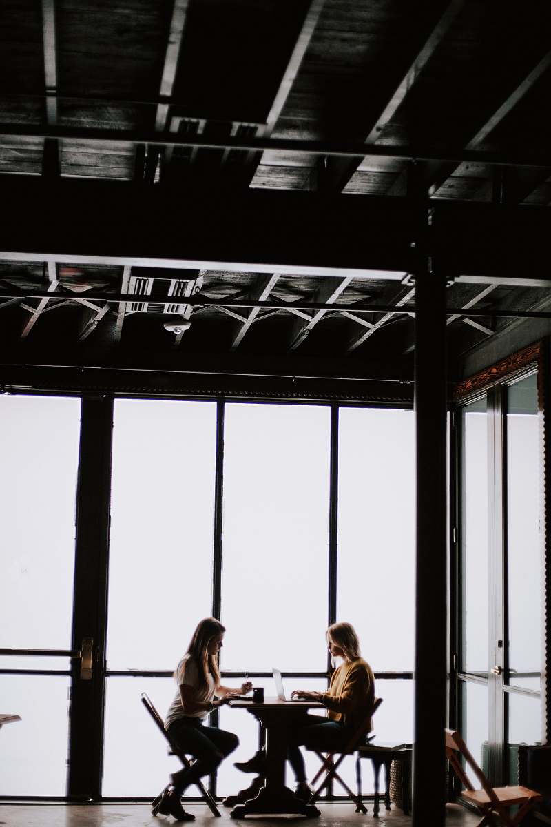 native hostel and bar & kitchen photo of two women facing each other sitting in front of table near glass wall austin